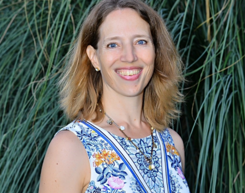 Photo of Julia from the shoulders up, standing in front of long green grass. She has shoulder length dark blonde hair and wears a blue paisley pattered vest top.