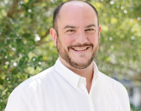 Photo of Mark standing in front of a green background with trees, smiling. He wears a white shirt.