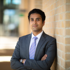 Akshay Mangla standing in the SBS hallway wearing a grey suit and smiling. 