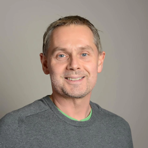 Alex Nicholls standing in front of a grey background and smiling.