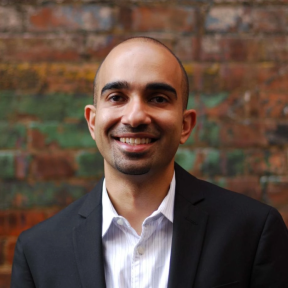 Karim Harji standing in front of a brick wall wearing a suit and smiling. 