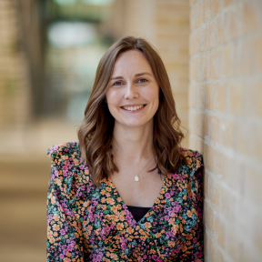 Lydia Allard standing in the SBS hallway wearing a floral top and smiling. 