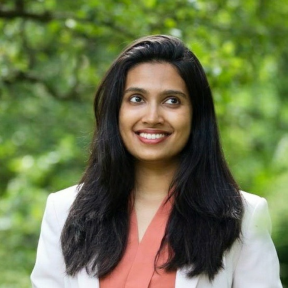 Sugandha Srivastav smiling standing in front a leafy and bright green background. 