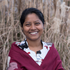 Anjali Sarker in front of a field smiling wearing a white patterned blouse with a red shawl.