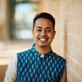 Avinash Kothuri smiling and standing in the SBS hallway wearing a blue patterned button-up and pale shirt.