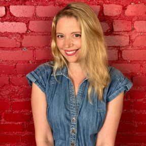 Charmian Love smiling in front of a red painted brick wall wearing a denim jumpsuit.