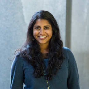 Christie George smiling in front of a grey background wearing a blue sweater.