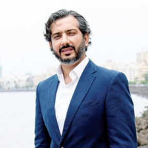 Dhruv Lakra standing in front of the sea in a pink shirt and blue blazer.