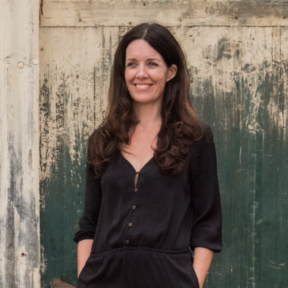 Emma Caddy standing in front of a wooden panel in a dark grey blouse, smiling.