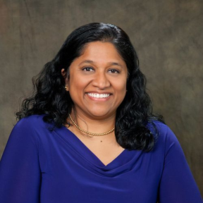 Esther Benjamin smiling in a royal blue dress and standing against a grey background.