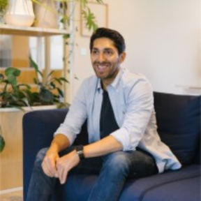 Faisal Butt sitting on a blue sofa with plants behind. He is smiling and wearing a pale blue shirt and jeans. 