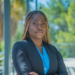 Folasade Ayoola is pictured standing with her arms crossed in front of a blurred natural background, wearing a blue shirt and black blazer.