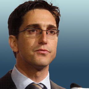 François Bonnici  in front of a blue background wearing a beige suit and tie.