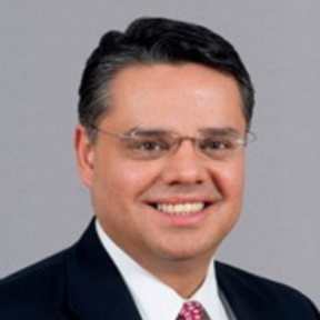 Henry Gonzalez standing in front of a grey background wearing a suit and red tie.