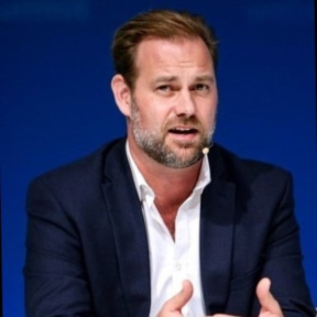 Jesse Moore sitting in a black blazer and white shirt talking in front of a blue background.