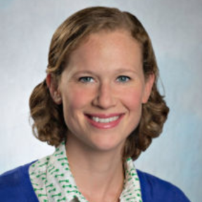 Lara Vogel smiling in front of a pale blue background wearing a blue cardigan and white shirt.