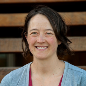 Lindsay Miller standing and smiling in front of wooden panels wearing a blue cardigan.