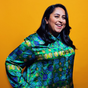 Maryam Pasha standing in front of a bright mustard yellow background, laughing and wearing a bright blue and emerald patterned shirt.