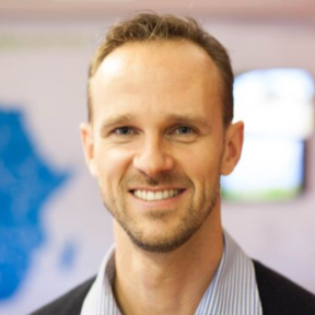 Mike Quinn smiling in front of a blurred background wearing a blue shirt.