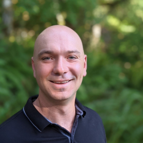 Nigel Tunnacliff standing in front of a leafy background smiling. 
