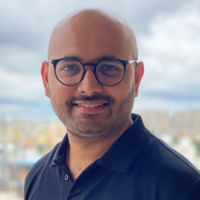 Nikhil Nair standing in front of a blurry city smiling wearing a branded t-shirt. 