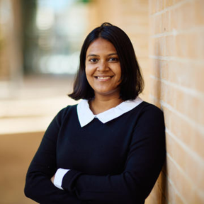 Nitya Nangalia leaning against a brick wall in the SBS courtyard, smiling and wearing a black sweater with white shirt. 