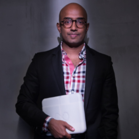 Rangan Srikhanta is pictured standing in a spotlight holding a laptop, wearing a blue and red checked shirt.
