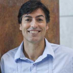 Richard Webb smiling in front of a wooden panel in a light blue shirt.