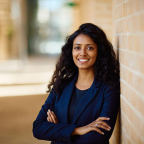 Shruti Jain leaning against a brick wall in the Said Business School courtyard, smiling and wearing a black blouse and navy blue blazer.