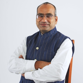 Sujeet Kumar sitting with his arms crossed in front of a white background wearing a pin stripe sleeveless button-up and white shirt.  