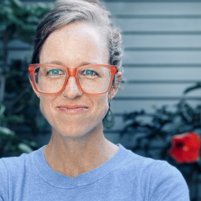 Sydney Schaef wearing bright red glasses and a blue top and smiling. 