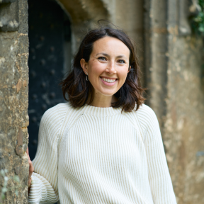 Jessica Jacobson standing in a college doorway wearing a white jumper and smiling. 