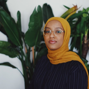 Izzy Mohammed standing in front of a green plant wearing a black top. 