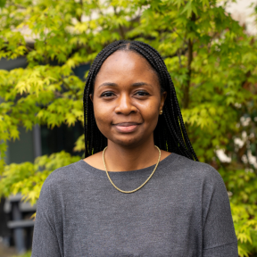 Aisha Raheem wearing a grey top in front of a green bush. 
