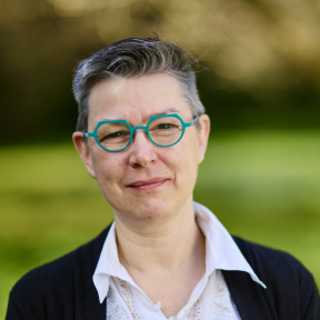 Close up headshot of Kathryn wearing a white shirt, black cardigan and circular turquoise-framed glasses. 