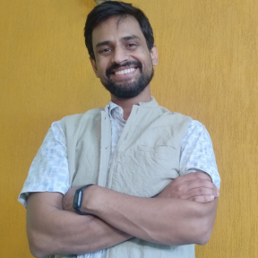 Saransh Vaswani is standing in front of a yellow wall with his arm crossed. He is smiling and wearing a white top. 