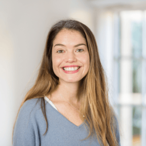 Headshot from shoulders up of Hayley grinning, in a well lit room. She is wearing a grey blue v neck jumper. 