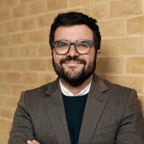 Headshot from shoulders up of Ignacio smiling in front of a yellow brick wall. He wears thick rimmed glasses, and a brown suit jacket with white shirt and dark brown jumper underneath. 