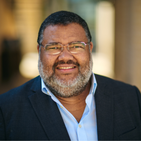 Athol Williams Headshot of Athol wearing a pale blue shirt and dark coloured blazer, smiling in front of a bright sunny background.