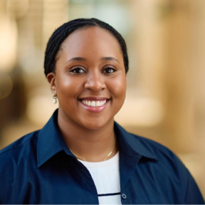 Rehema Msulwa Headshot from the shoulders up of Rehema smiling in front of a bright sunny background. She is wearing a dark blue shirt and a white top with a black horizontal stripe across.