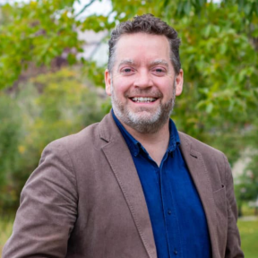 James Stauch is smiling in front of greenery, wearing a brown jacket and a blue shirt. 