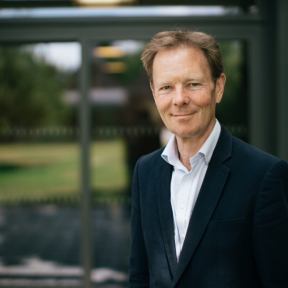 Jonathon is wearing a dark suit and smiling in front of a bright window. 