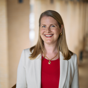 Mary is wearing a cream blazer and a red top. She is standing in a hallway, smiling. 