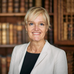 Mette is wearing a cream jacket and is smiling in front of a bookcase.