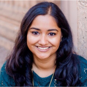 Shivani in standing in front of a brick wall and smiling wearing a blue top. 