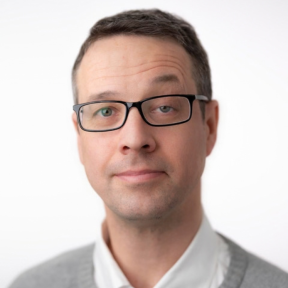 Headshot of Howard. He wears glasses, has short dark hair and is wearing a white shirt with grey blazer. 