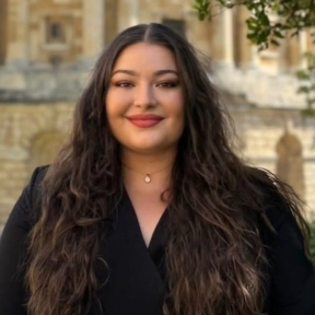 Headshot of Jasmine with an Oxford building in the background. She has long brown hair and is wearing a black top.