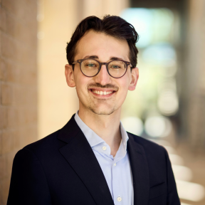 Headshot of Lukas in a warmly lit hallway. He wears round glasses, is smiling and wears a black blazer and pale blue shirt. 