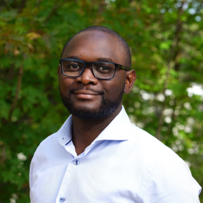 Headshot of Maganizo in front of a green hedge. He wears glasses, has very short hair and is wearing a white shirt. 