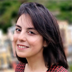Heasdshot of Vira in front of a green/urban blurred background. She has long dark hair and is wearing a black and red checkered top. 